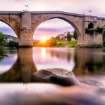 Old stone bridge over calm river at sunrise, symbolising enduring foundations and quiet strength.