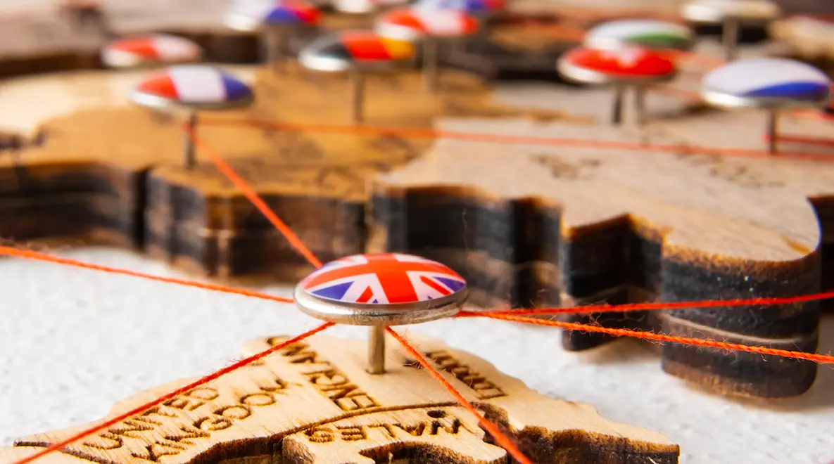 Pin with UK flag connected by red threads on a wooden map of Europe.